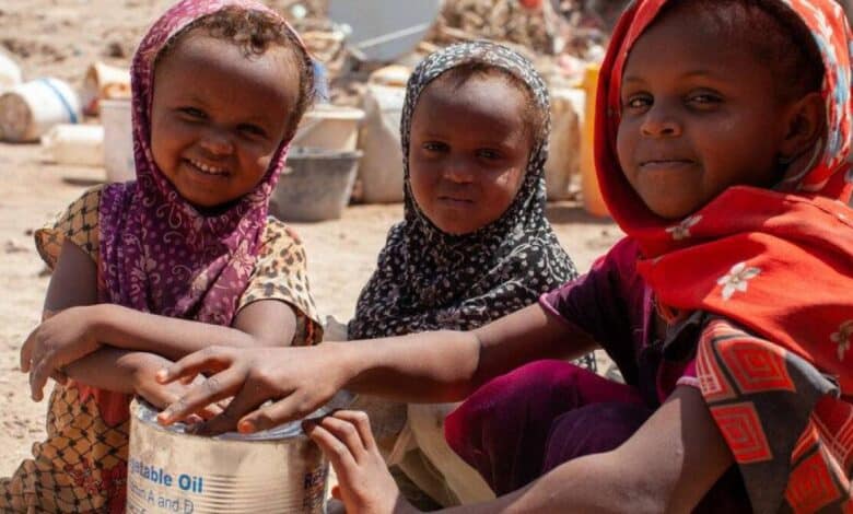 The image of a three children in colourful headscarves sit together, holding a tin of vegetable oil labelled with the WFP (World Food Programme) logo, in a dusty area with barrels and containers in the background.
