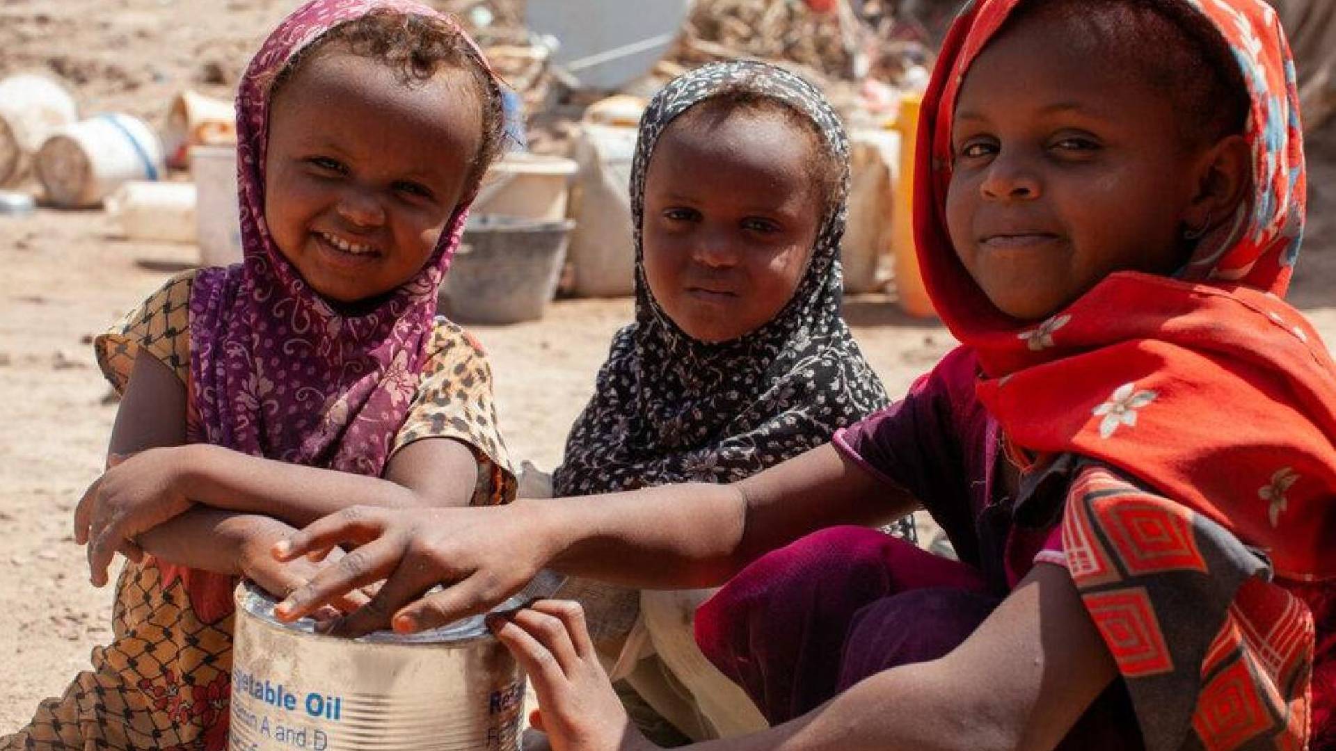 The image of a three children in colourful headscarves sit together, holding a tin of vegetable oil labelled with the WFP (World Food Programme) logo, in a dusty area with barrels and containers in the background.