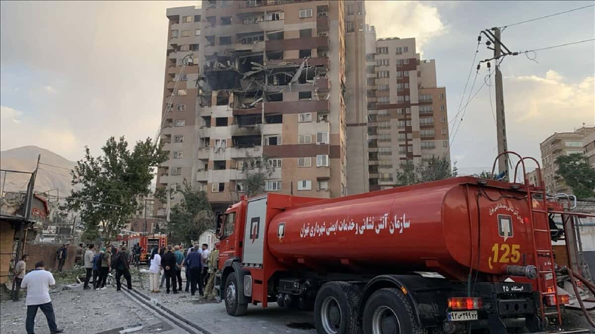 A damaged building in Tehran, Iran, with visible fire and structural collapse, surrounded by emergency response vehicles and personnel, set against a backdrop of trees and mountains, captured on June 13, 2025.