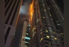 Flames and smoke rise from the upper floors of a high-rise residential tower at night in Dubai Marina, with neighbouring buildings visible in the foreground.