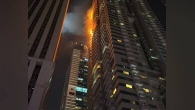 Flames and smoke rise from the upper floors of a high-rise residential tower at night in Dubai Marina, with neighbouring buildings visible in the foreground.