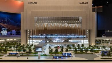 A detailed architectural model of the Emaar Properties Station on the Dubai Metro Blue Line, showcasing a futuristic and illuminated structure with pedestrian bridges, greenery, and buses in the foreground. The tall, modern design features vertical lighting and a grand facade.