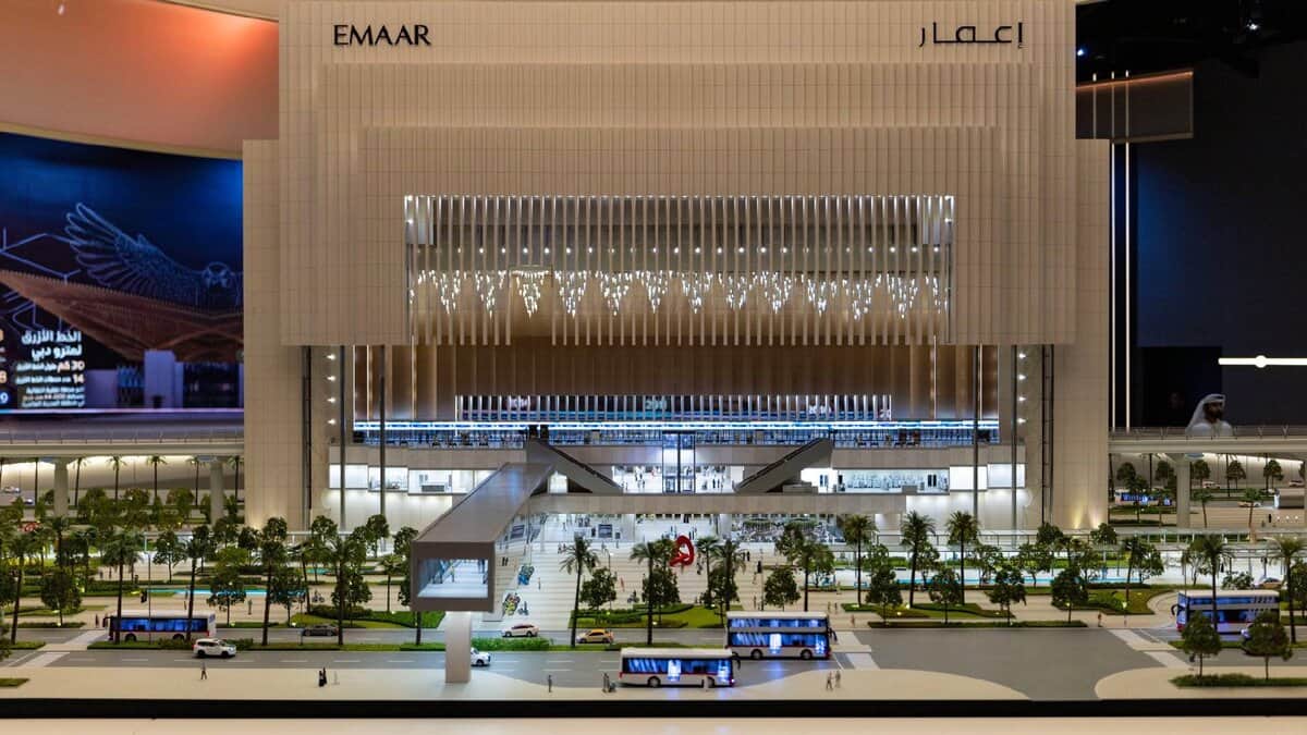 A detailed architectural model of the Emaar Properties Station on the Dubai Metro Blue Line, showcasing a futuristic and illuminated structure with pedestrian bridges, greenery, and buses in the foreground. The tall, modern design features vertical lighting and a grand facade.