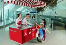 Two Emirates staff serve complimentary ice cream from a red and white striped cart to two young children at Dubai International Airport, with a modern terminal and travelators in the background.