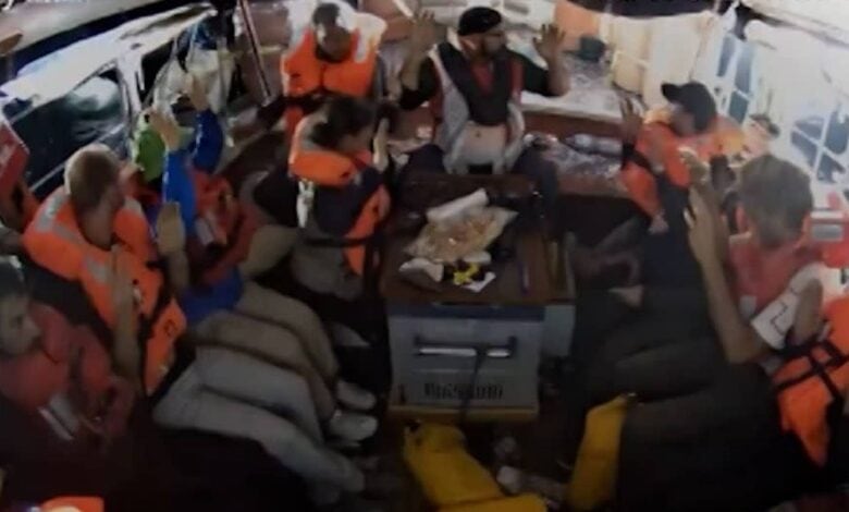 Crew members of a Gaza-bound aid ship wearing life jackets sit closely together with hands raised inside the vessel after being detained by Israeli forces.