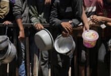 A group of Palestinians, mostly men and boys, stand in line holding empty pots and containers, waiting for food aid behind a metal barrier in Gaza.