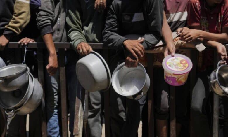 A group of Palestinians, mostly men and boys, stand in line holding empty pots and containers, waiting for food aid behind a metal barrier in Gaza.