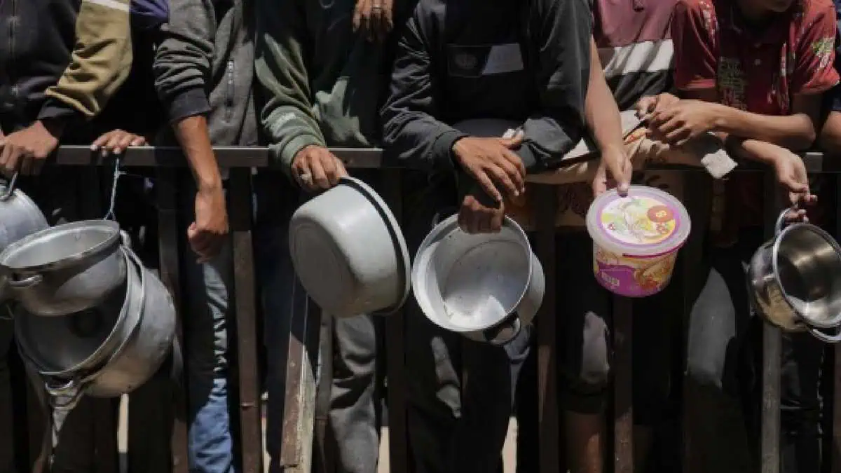 A group of Palestinians, mostly men and boys, stand in line holding empty pots and containers, waiting for food aid behind a metal barrier in Gaza.