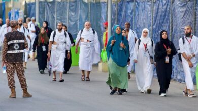 Pilgrims walk towards Mina on the morning of the Day of Tarwiyah during Haj 2025, escorted by Saudi security personnel. Men are dressed in white ihram, while women wear modest attire with ID lanyards visible.
