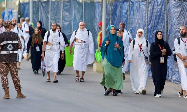 Pilgrims walk towards Mina on the morning of the Day of Tarwiyah during Haj 2025, escorted by Saudi security personnel. Men are dressed in white ihram, while women wear modest attire with ID lanyards visible.