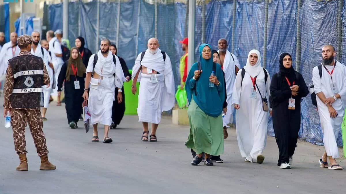 Pilgrims walk towards Mina on the morning of the Day of Tarwiyah during Haj 2025, escorted by Saudi security personnel. Men are dressed in white ihram, while women wear modest attire with ID lanyards visible.