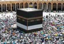 A large crowd of Haj pilgrims, many holding colourful umbrellas, perform the farewell Tawaf around the Kaaba at the Grand Mosque in Makkah, under bright sunlight.