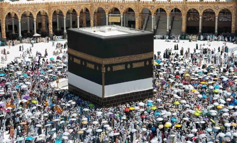 A large crowd of Haj pilgrims, many holding colourful umbrellas, perform the farewell Tawaf around the Kaaba at the Grand Mosque in Makkah, under bright sunlight.