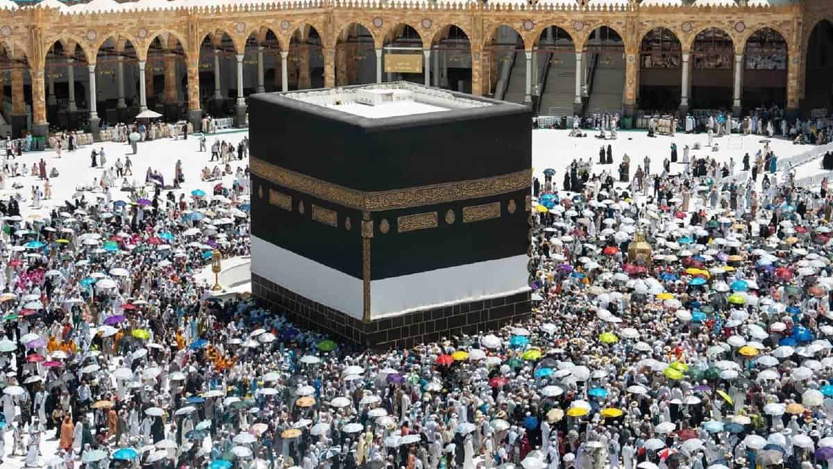 A large crowd of Haj pilgrims, many holding colourful umbrellas, perform the farewell Tawaf around the Kaaba at the Grand Mosque in Makkah, under bright sunlight.