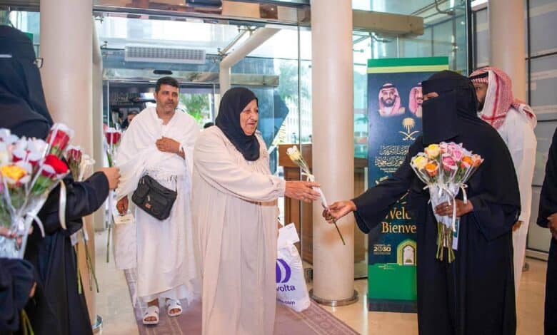 A female pilgrim receives a flower from a Saudi official as she arrives for Haj, with other pilgrims following behind in a warmly decorated arrival area.