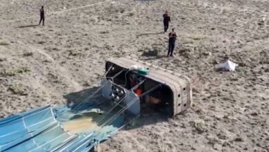 The image of a toppled hot air balloon basket lies on its side in a dry, rocky field in central Turkey. The balloon’s fabric is deflated on the ground, and several emergency responders are seen near the crash site, with a police vehicle and cordoned-off area in the background.