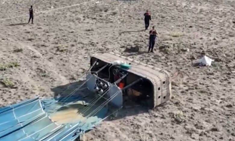 The image of a toppled hot air balloon basket lies on its side in a dry, rocky field in central Turkey. The balloon’s fabric is deflated on the ground, and several emergency responders are seen near the crash site, with a police vehicle and cordoned-off area in the background.