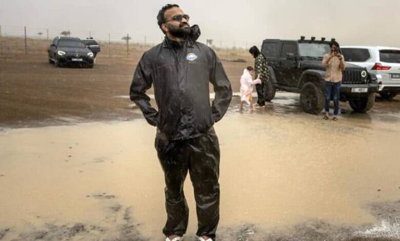 Muhammed Sajjad, known locally as “The Weatherman”, stands in a puddle wearing a black raincoat and flip-flops during rain in the Sharjah desert, with off-road vehicles and other participants in the background.