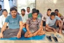 The image of a group of Indian migrant workers sit on mats and cardboard sheets against a wall, some looking directly at the camera while others appear visibly distressed.