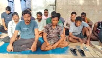 The image of a group of Indian migrant workers sit on mats and cardboard sheets against a wall, some looking directly at the camera while others appear visibly distressed.