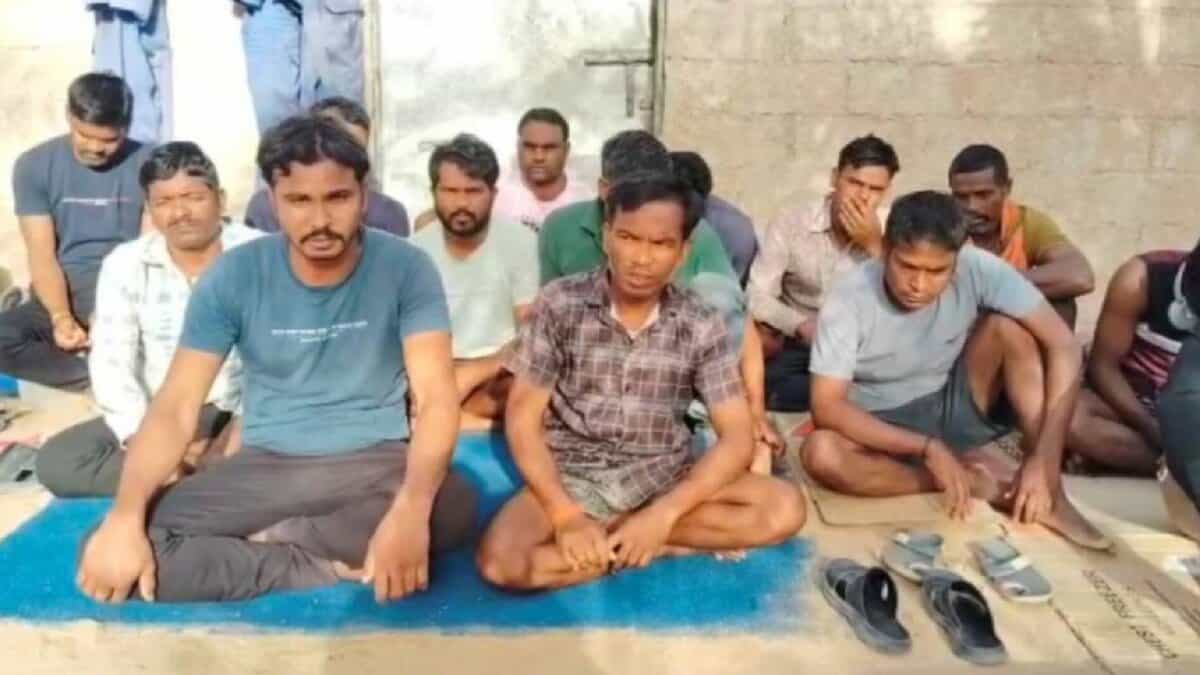 The image of a group of Indian migrant workers sit on mats and cardboard sheets against a wall, some looking directly at the camera while others appear visibly distressed.