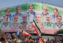 A large crowd gathers beneath a massive mural in Tehran showing illustrated portraits of senior Iranian military commanders and nuclear scientists who were killed in recent Israeli airstrikes. The background of the mural features the Iranian flag, with the word “شهید” (martyr) visible in Persian script. People in the crowd are holding Iranian flags and banners.