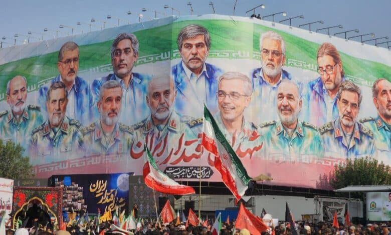 A large crowd gathers beneath a massive mural in Tehran showing illustrated portraits of senior Iranian military commanders and nuclear scientists who were killed in recent Israeli airstrikes. The background of the mural features the Iranian flag, with the word “شهید” (martyr) visible in Persian script. People in the crowd are holding Iranian flags and banners.