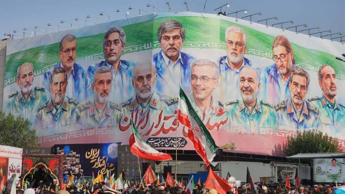 A large crowd gathers beneath a massive mural in Tehran showing illustrated portraits of senior Iranian military commanders and nuclear scientists who were killed in recent Israeli airstrikes. The background of the mural features the Iranian flag, with the word “شهید” (martyr) visible in Persian script. People in the crowd are holding Iranian flags and banners.