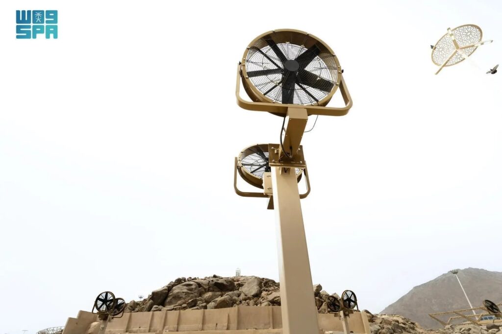 A tall structure with large industrial fans is installed on a rocky terrain at Jabal Al-Rahmah, with mountains in the background. The Kidana logo is visible in the top left corner, and a decorative element with birds is seen in the sky.