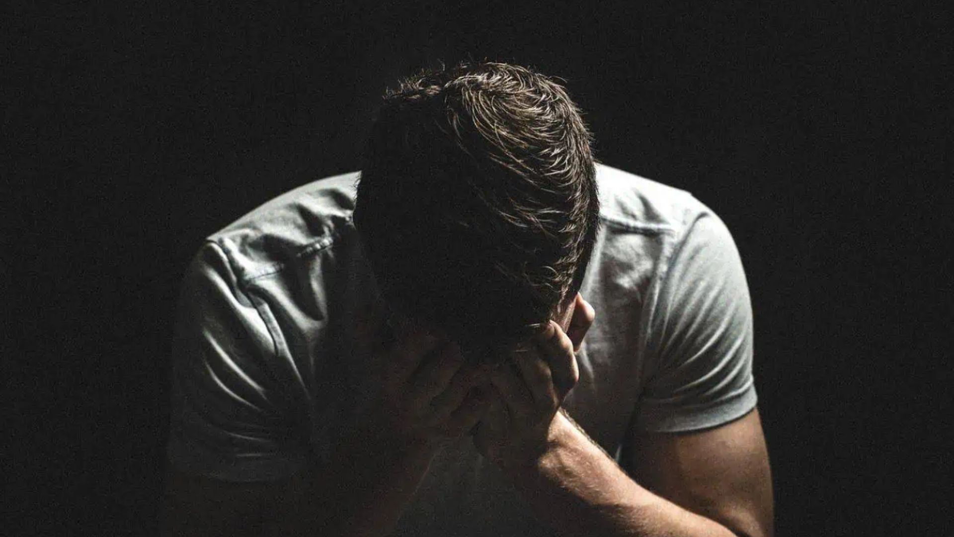 A shadowy image of a man with his head bowed and hands covering his face, wearing a gray shirt. The dark background emphasizes a mood of despair or deep thought, creating a somber and introspective atmosphere.