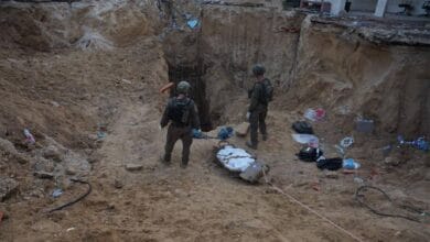 Two Israeli soldiers stand at the edge of a deep sandy excavation site near an underground tunnel in Khan Younis, southern Gaza. A body wrapped in a white shroud lies on a stretcher, being pulled out from the tunnel amid scattered belongings and debris.