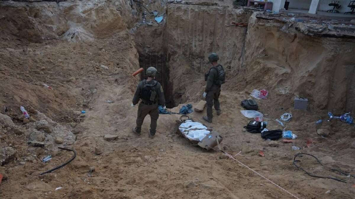 Two Israeli soldiers stand at the edge of a deep sandy excavation site near an underground tunnel in Khan Younis, southern Gaza. A body wrapped in a white shroud lies on a stretcher, being pulled out from the tunnel amid scattered belongings and debris.