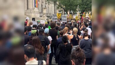 Large crowd gathered outside the New York Public Library during an anti-war rally, with placards reading “HANDS OFF IRAN” and “END ALL U.S. AID TO ISRAEL” visible above the crowd. Trees and skyscrapers frame the scene.