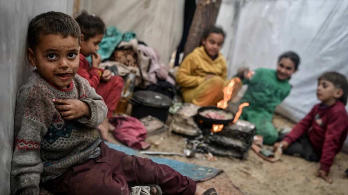 A group of Palestinian children in worn clothing sit inside a makeshift tent, with one child in the foreground looking at the camera, their face and jumper smeared with dirt. In the background, children gather around a small open fire cooking food in a pan.