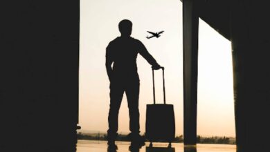 The image of a silhouette of a man standing at an airport terminal, holding a suitcase, with an aeroplane visible in the sky outside at sunset.