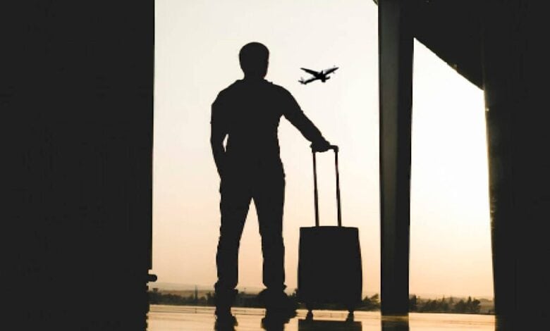 The image of a silhouette of a man standing at an airport terminal, holding a suitcase, with an aeroplane visible in the sky outside at sunset.
