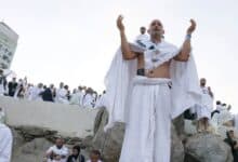 The image of Haj pilgrims gather on Mount Arafat in Makkah on the second day of Haj 2025. A person stands on a large rock with arms raised in prayer, while others sit and stand around, some taking photos. A white structure with Arabic text is visible in the background. The scene is set on a rocky, open terrain under a clear sky.
