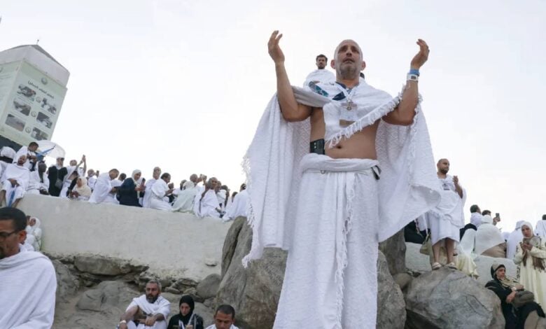 The image of Haj pilgrims gather on Mount Arafat in Makkah on the second day of Haj 2025. A person stands on a large rock with arms raised in prayer, while others sit and stand around, some taking photos. A white structure with Arabic text is visible in the background. The scene is set on a rocky, open terrain under a clear sky.