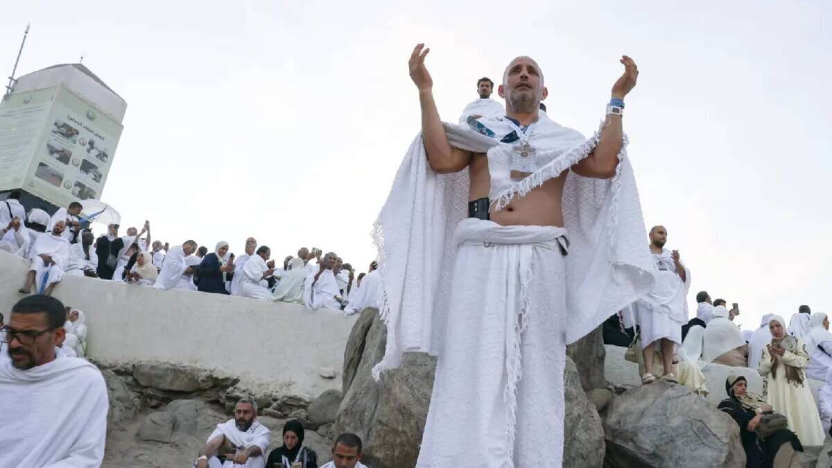 The image of Haj pilgrims gather on Mount Arafat in Makkah on the second day of Haj 2025. A person stands on a large rock with arms raised in prayer, while others sit and stand around, some taking photos. A white structure with Arabic text is visible in the background. The scene is set on a rocky, open terrain under a clear sky.