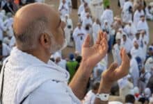 A Haj pilgrim in white ihram raises his hands in prayer, surrounded by a large crowd of fellow worshippers at Mount Arafat.