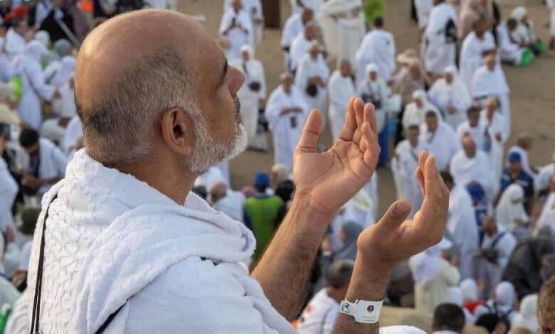 A Haj pilgrim in white ihram raises his hands in prayer, surrounded by a large crowd of fellow worshippers at Mount Arafat.