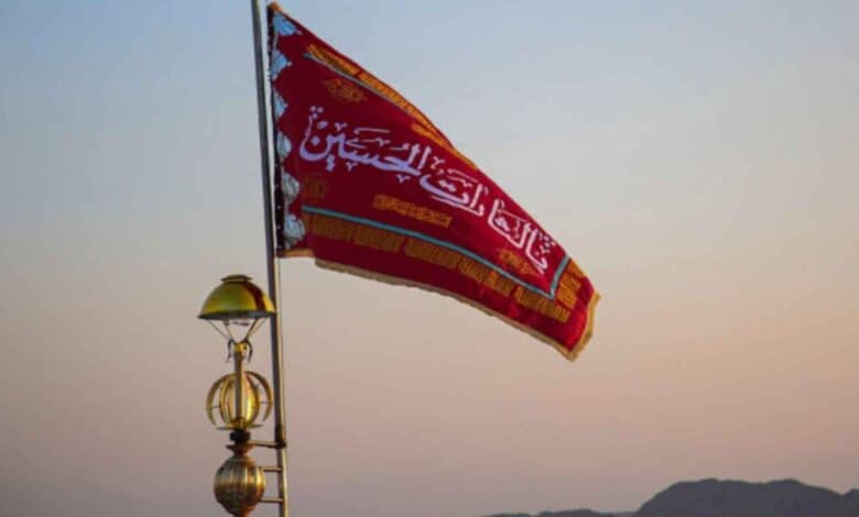 A red flag with Arabic calligraphy is hoisted on a pole against a dusky sky, symbolising mourning and a call for vengeance in Shiite tradition.