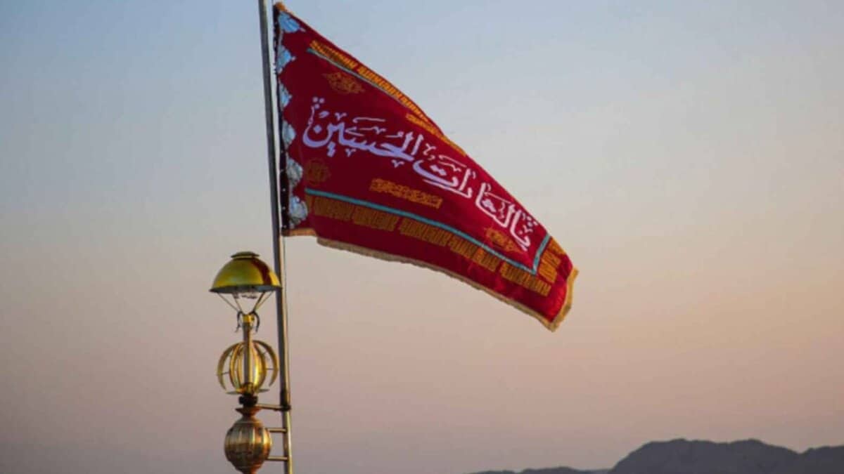 A red flag with Arabic calligraphy is hoisted on a pole against a dusky sky, symbolising mourning and a call for vengeance in Shiite tradition.