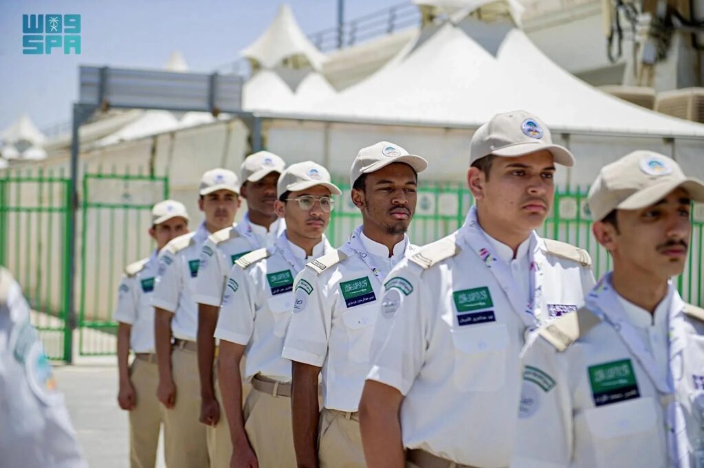A group of scouts in white uniforms and beige caps stand in a line outdoors, with white tents and green fencing in the background.
