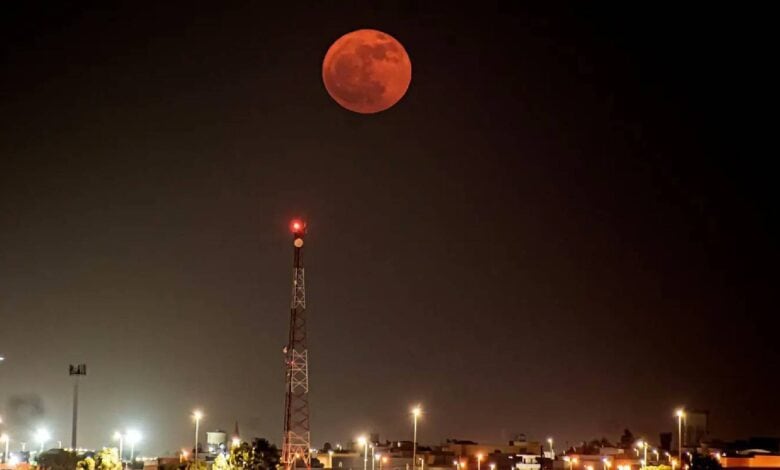 A vivid reddish full moon, known as the Strawberry Moon, glows brightly in the night sky above a cityscape in Saudi Arabia, with a lit communication tower and buildings below under artificial lights.