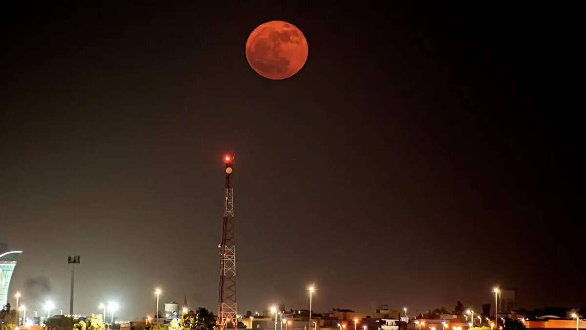 A vivid reddish full moon, known as the Strawberry Moon, glows brightly in the night sky above a cityscape in Saudi Arabia, with a lit communication tower and buildings below under artificial lights.