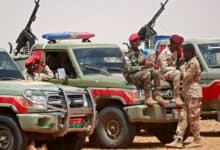 The image of Sudanese paramilitary Rapid Support Forces (RSF) soldiers in uniform sit and stand around armed pickup trucks in a desert area, equipped with mounted machine guns.