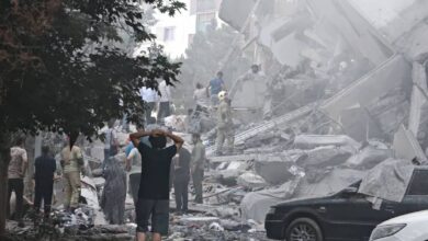 People search through rubble and damaged vehicles after a building collapsed in Tehran, Iran. The area is filled with dust and debris as rescuers and civilians gather at the scene of destruction caused by airstrikes.