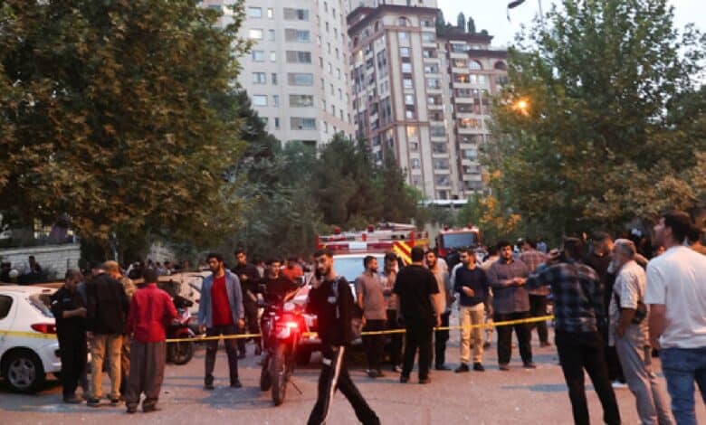 The image of a crowd gathers on a Tehran street amidst emergency vehicles and police tape, following Israeli strikes, with tall buildings and trees in the background, on June 13, 2025.