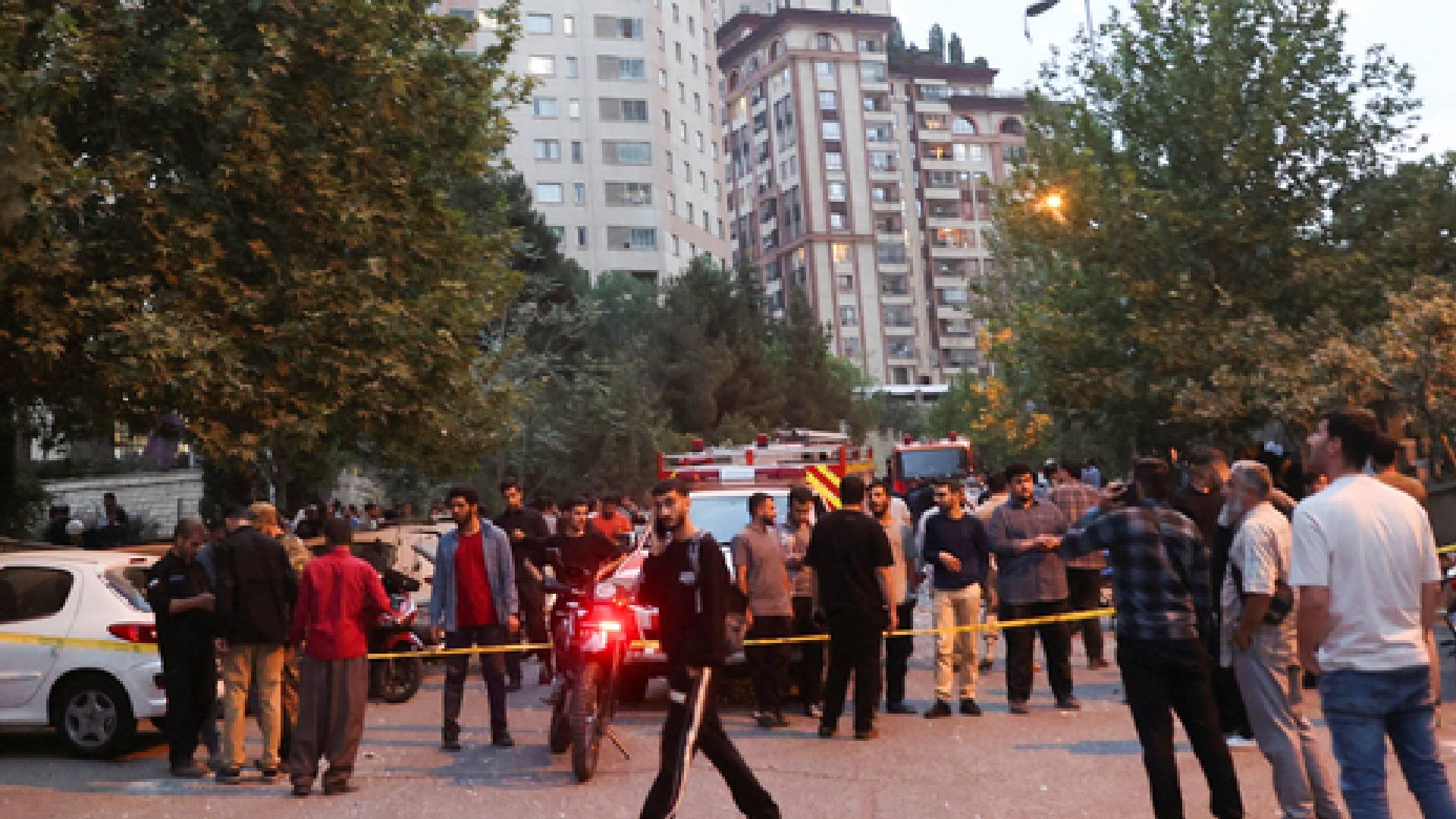 The image of a crowd gathers on a Tehran street amidst emergency vehicles and police tape, following Israeli strikes, with tall buildings and trees in the background, on June 13, 2025.